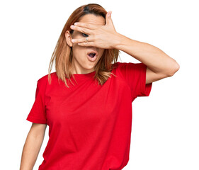 Hispanic young woman wearing casual red t shirt peeking in shock covering face and eyes with hand, looking through fingers with embarrassed expression.
