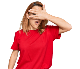 Hispanic young woman wearing casual red t shirt peeking in shock covering face and eyes with hand, looking through fingers with embarrassed expression.