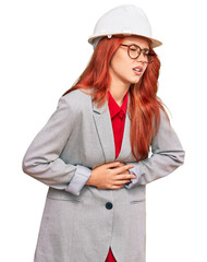 Young redhead woman wearing architect hardhat with hand on stomach because nausea, painful disease feeling unwell. ache concept.