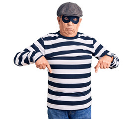 Senior handsome man wearing burglar mask and t-shirt pointing down looking sad and upset,...