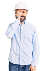 Young handsome man wearing architect hardhat covering one eye with hand, confident smile on face and surprise emotion.