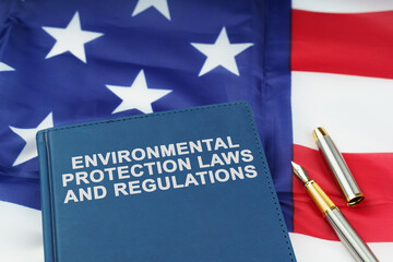 On the US flag lies a pen and a book with the inscription - ENVIRONMENTAL PROTECTION LAWS AND REGULATIONS - obrazy, fototapety, plakaty