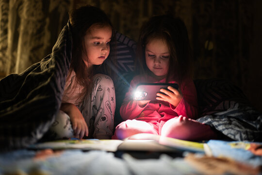 Two sisters watching movie or cartoon in smartphone together with blanket cover in bed at night home.