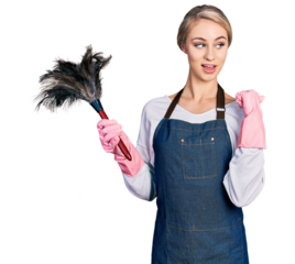 Beautiful young blonde woman wearing apron holding cleaning duster smiling with happy face looking and pointing to the side with thumb up.