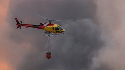 Fire brigade helicopter trying to put out a fire with water, surrounded by smoke and burning trees.