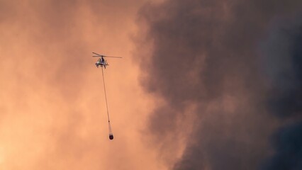 Fire brigade helicopter trying to put out a fire with water, surrounded by smoke and burning trees.