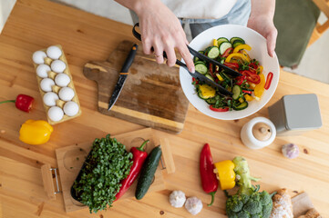 Top view of hands of a young woman preparing a salad of fresh vegetables and greens in a white bowl, mixing it with salad tongs over a wooden table. Healthy Eating. Horizontal photo.