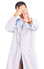 Handsome young man with bear wearing doctor uniform covering eyes with hands and doing stop gesture with sad and fear expression. embarrassed and negative concept.