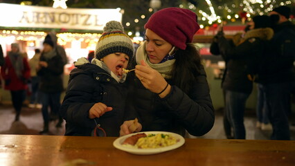 Mother and child eating food in winter Christmas market in Europe. Mom feeding little boy food seated at table during festivities