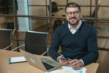 Smiling man using smartphone and working with laptop in the office