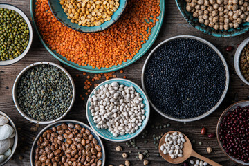 Different legumes (lentils, beans, chickpeas, mung, peas) in bowls and plates on wooden background.