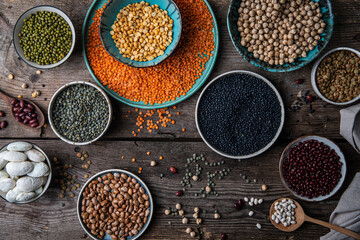 Different legumes (lentils, beans, chickpeas, mung, peas) in bowls and plates on wooden background.