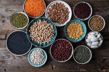 Different legumes (lentils, beans, chickpeas, mung, peas) in bowls and plates on wooden background.