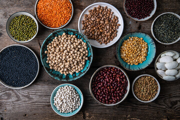 Different legumes (lentils, beans, chickpeas, mung, peas) in bowls and plates on wooden background.