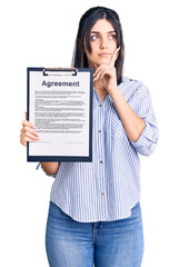 Young beautiful girl holding clipboard with agreement document serious face thinking about question with hand on chin, thoughtful about confusing idea