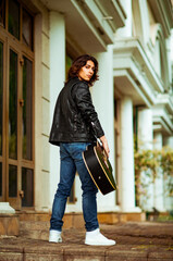 A young, handsome man with long hair and a black guitar, on the streets of the city