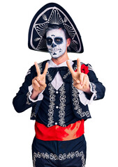 Young man wearing day of the dead costume over background smiling looking to the camera showing fingers doing victory sign. number two.