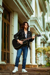 A young, handsome man with long hair and a black guitar, on the streets of the city