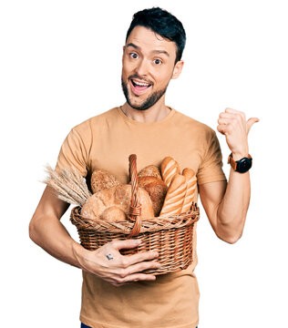 Hispanic man with beard holding wicker basket with bread pointing thumb up to the side smiling happy with open mouth