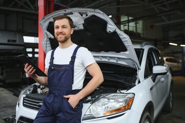 Bearded mechanic in overalls standing in garage of a car salon and holding tablet. He is about to diagnostic breakdown.