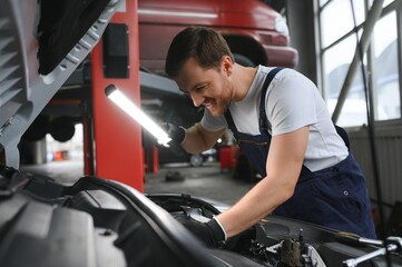 Handsome young male auto mechanic in special uniform clothes holding a flashlight, looking for breakdown and repairing under the hood in the car engine in a car workshop