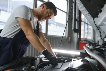 Handsome young male auto mechanic in special uniform clothes holding a flashlight, looking for breakdown and repairing under the hood in the car engine in a car workshop