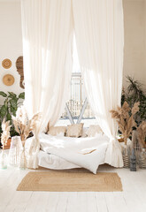 Bed with pillows, wooden furniture, plants in pots, an armchair and curtains on large windows in a...