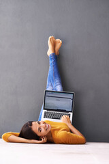 The up-side of technology. A young woman lying on the floor with her laptop against a gray...