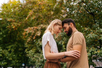 Low-angle view of happy young couple in love standing with closed eyes in front of each other at city park on background of trees. Tattooed handsome man and pretty lady walking spending time together.