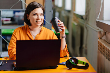 a brunette woman in an orange blouse drinks coffee and works on a laptop