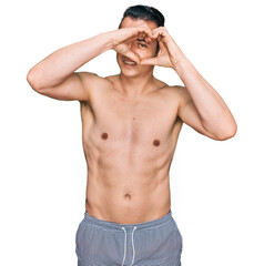 Fototapeta premium Handsome young man wearing swimwear shirtless doing heart shape with hand and fingers smiling looking through sign