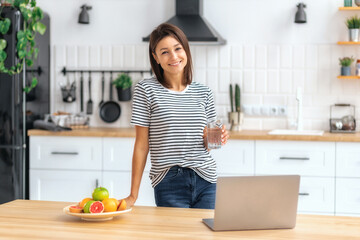 Portrait of beautiful woman holding glass of water standing in the kitchen at home looking at the camera and smiling, wellness healthy food concept