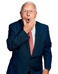 Senior man with grey hair wearing business suit and glasses looking fascinated with disbelief, surprise and amazed expression with hands on chin