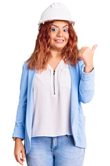 Young latin woman wearing architect hardhat smiling with happy face looking and pointing to the side with thumb up.