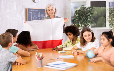 Fototapeta na wymiar Mature primary school teacher working in a high school tells pupils the history of Poland in class and holds the national flag .of the country in her hands