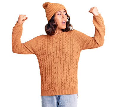 Young beautiful mixed race woman wearing wool sweater and winter hat showing arms muscles smiling proud. fitness concept.