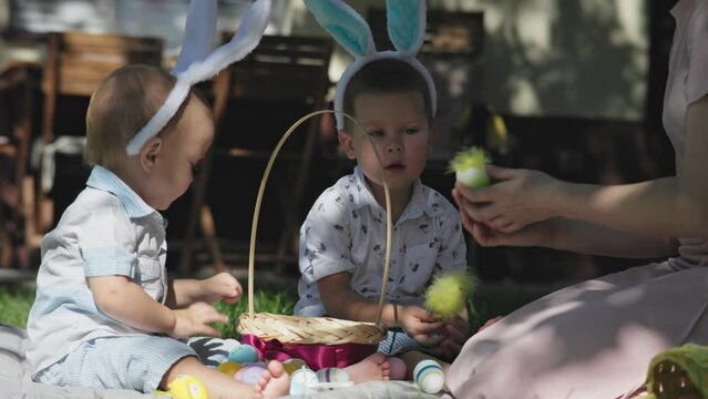 happy family celebrating Easter holiday outdoors. mother and little kids boys bunny ears sitting grass backyard having traditional Easter picnic