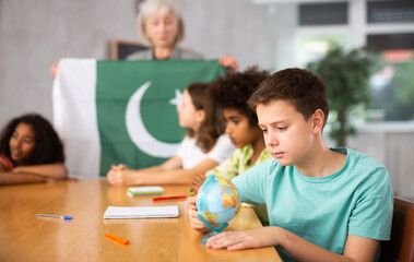 Fototapeta premium Mature woman teacher tells the pupils the history of Pakistan in the lesson and holds the national flag of the country
