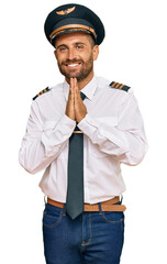Handsome man with beard wearing airplane pilot uniform praying with hands together asking for forgiveness smiling confident.