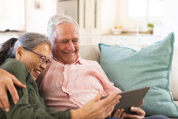 Happy senior diverse couple sitting on sofa and using tablet