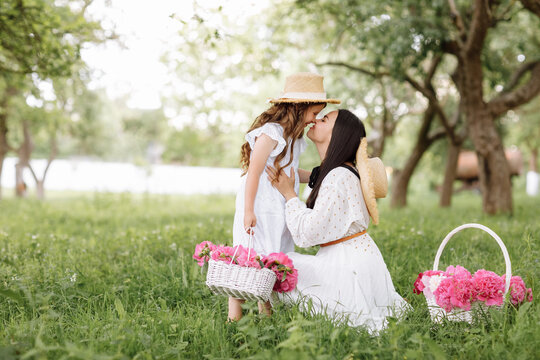 Mothers Day, Womens day. Young beautiful mother is spending time with little daughter in the green summer park. Mom with child are holding baskets of peonies flowers together on nature. Family look