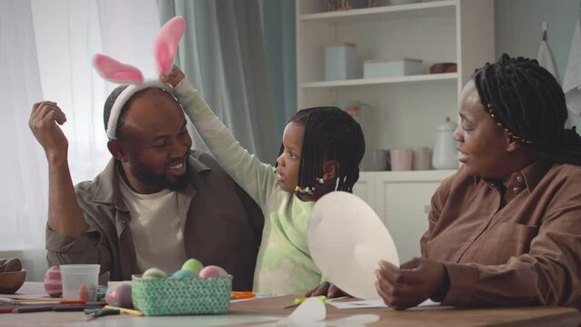 Medium shot of happy African American gamily of three celebrating Easter holiday together at home. Little girl putting on funny bunny ears on father head