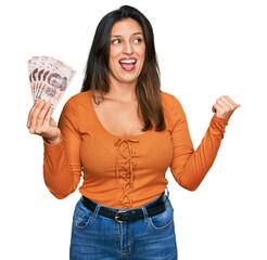 Obraz premium Beautiful hispanic woman holding mexican pesos pointing thumb up to the side smiling happy with open mouth