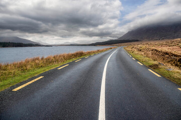Beautiful scenery and small road by a lake in Connemara, Ireland. Mountains and cloudy sky in the background. Travel and transportation concept.