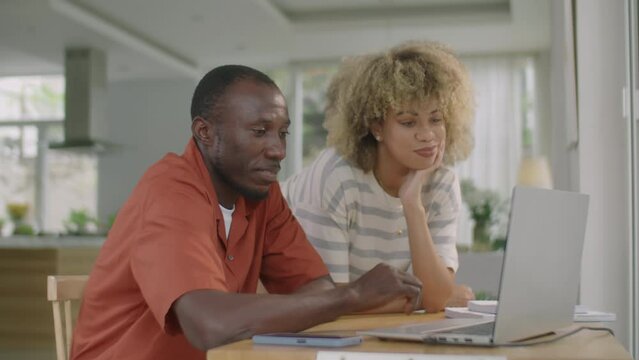 Medium shot of young diverse couple discussing something on laptop screen while doing online shopping at home