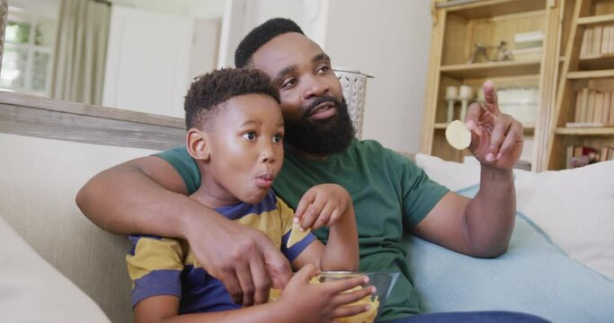 Happy african american father and son sitting on sofa, watching tv and eating crisps, in slow motion