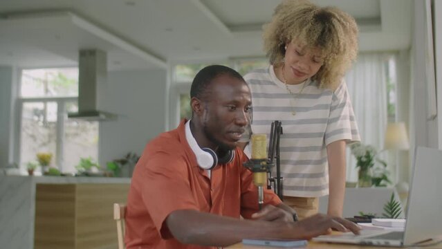 Medium shot of young diverse couple discussing something on laptop while recording music or podcast together in home studio