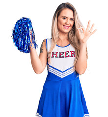 Young beautiful blonde woman wearing cheerleader uniform holding pompom doing ok sign with fingers,...
