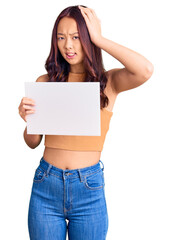 Young beautiful chinese girl holding paper banner with blank space stressed and frustrated with hand on head, surprised and angry face