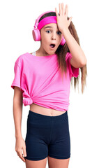 Cute hispanic child girl wearing gym clothes and using headphones surprised with hand on head for mistake, remember error. forgot, bad memory concept.